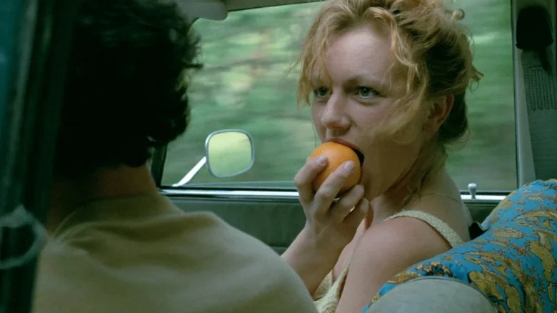 Johanna ter Steege biting into an orange fruit in passenger seat and looking at Gene Bervoets as Rex (face not pictured) in The Vanishing