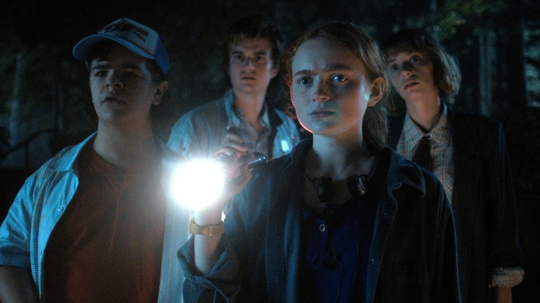 Gaten Matarazzo as Dustin and Sadie Sink as Max with a flashlight standing in front of Joe Keery as Steve and Maya Hawke as Robin in the woods in Stranger Things