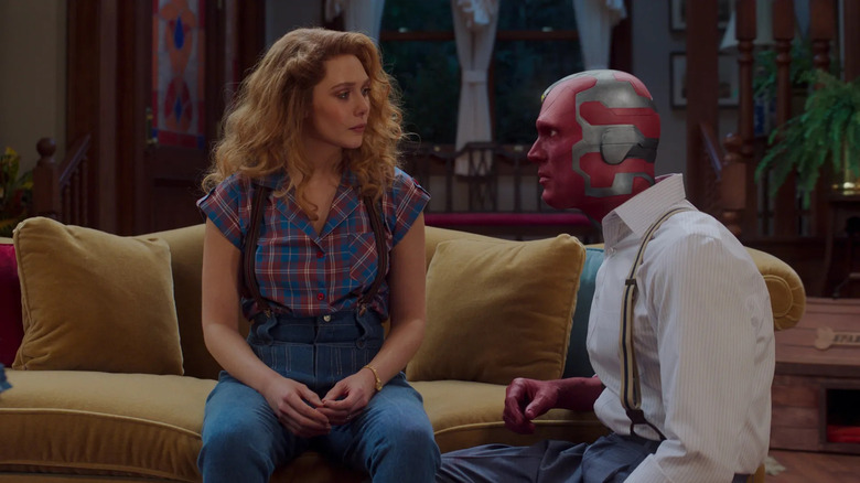 Elizabeth Olsen as Wanda sitting on the couch as Paul Bettany as Vision kneels next to her in WandaVision