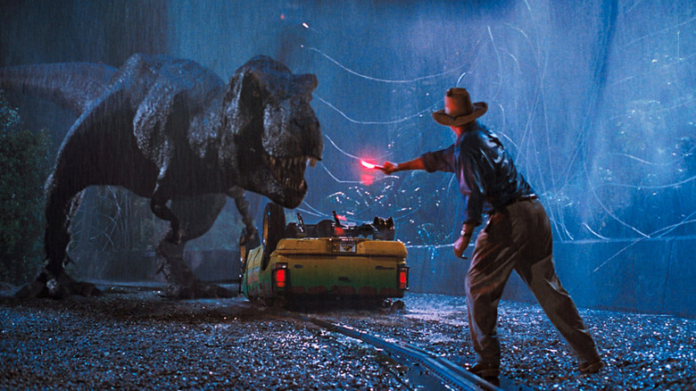 Sam Neill as Alan Grant in Jurassic Park, seen from behind as he waves a flare at a big, roaring T-Rex.