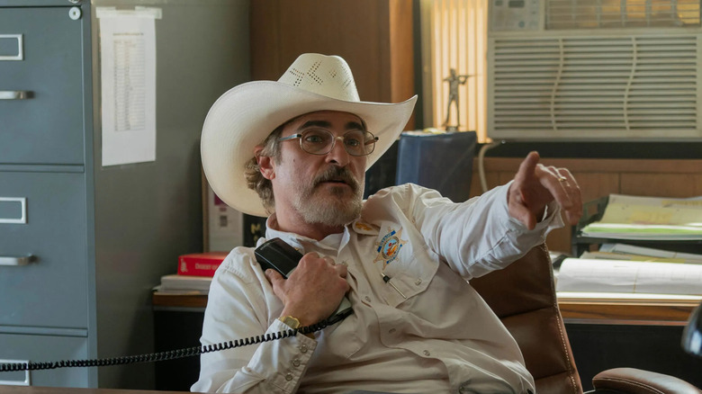 Joe sits at his desk and holds a phone in Eddington