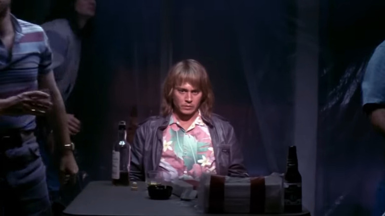 Johnny Depp with blonde hair and bangs in Hawaiian shirt sitting at table with bottles of alcohol on it in Blow