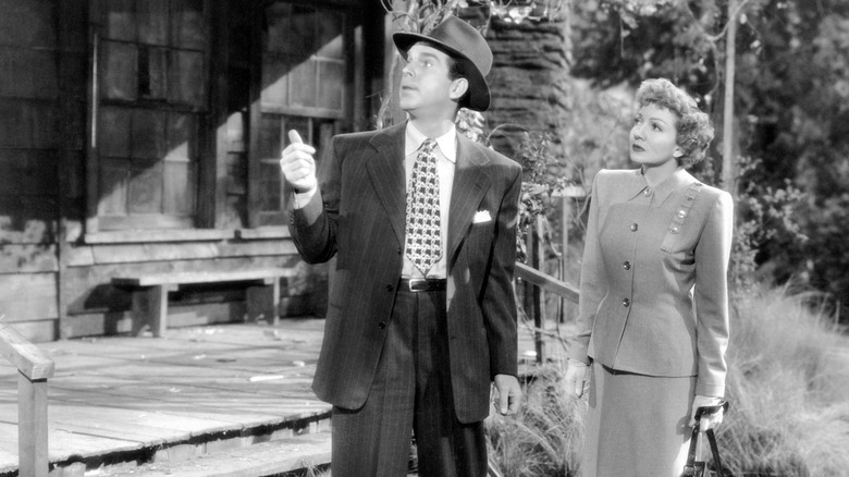 Fred MacMurray as Bob in suit and hat looking up at house next to Claudette Colbert as Betty in skirt suit in The Egg and I