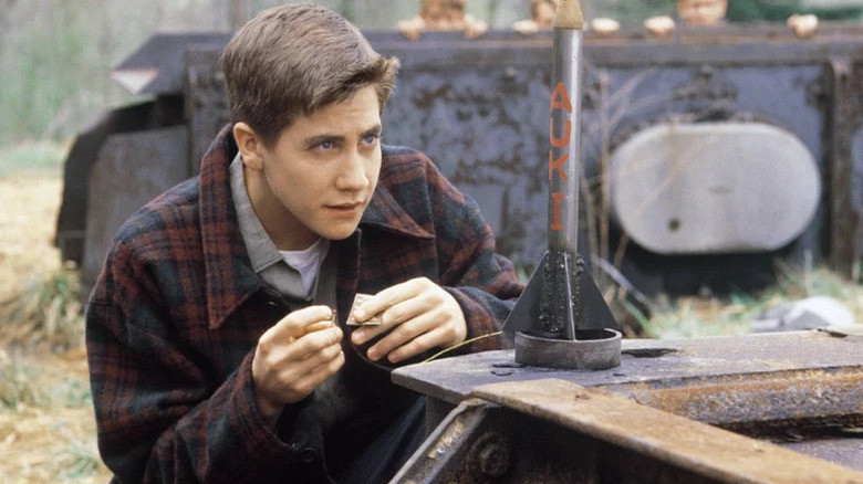 Jake Gyllenhaal as Homer in plaid jacket kneeling in front of small rocket in October Sky