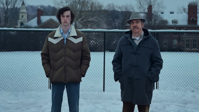 Dominic Sessa as Angus standing in snow in brown coat looking angry next to Paul Giamatti as Paul in blue coat and hat in The Holdovers