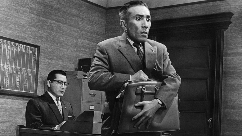 Nishi (Toshirô Mifune) looks angry at a desk behind Contract Officer Shirai (Kô Nishimura) worriedly holding a briefcase in The Bad Sleep Well