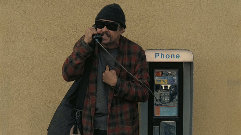Bob Ferguson talking on a payphone in a red bathrobe, beanie and sunglasses in One Battle After Another