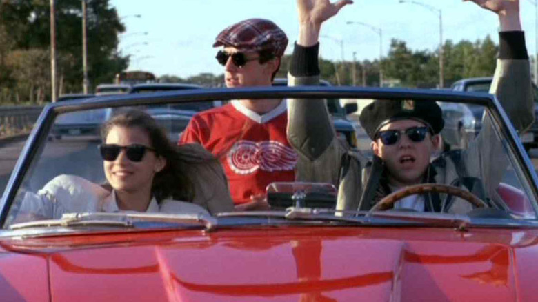 Matthew Broderick, Alan Ruck, and Mia Sara as Ferris, Cameron, and Sloane riding in the Ferrari in Ferris Bueller's Day Off, driving down the road in a convertible car, Ferris with his hands up.