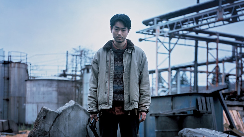 Yoshii stands amidst some factory ruins and holds a gun in Cloud