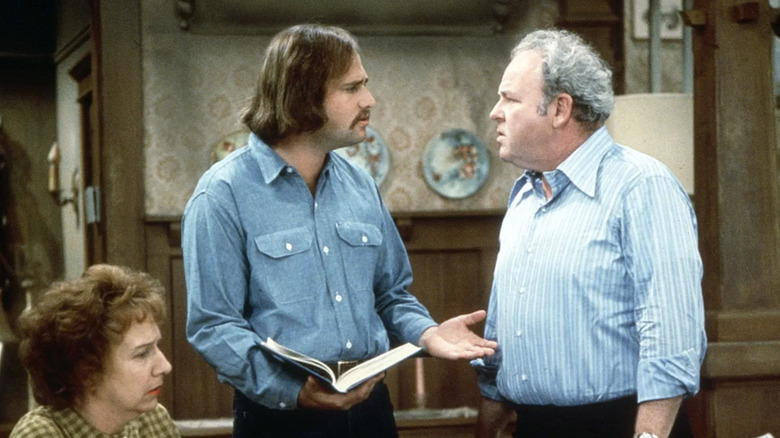 Jean Stapleton as Edith Bunker sitting while Rob Reiner as Michael "Meathead" Stivic holds a book and argues with Carroll O'Connor as Archie Bunker in All i the Family