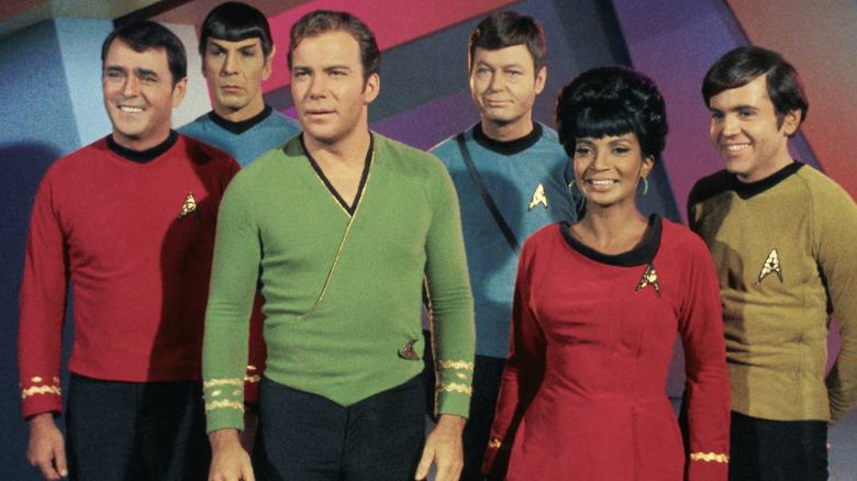 James Doohan as Montgomery "Scotty" Scott, Leonard Nimoy as Spock, William Shatner as Captain Kirk, DeForest Kelley as Leonard "Bones" McCoy, Nichelle Nichols as Nyota Uhura, and Walter Koenig as Pavel Chekov standing together in front of a colorful wall in Star Trek: The Original Series