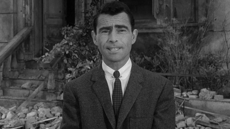Rod Serling hosting The Twiligjt Zone in front of a broken down house with bricks and dead plants on the ground