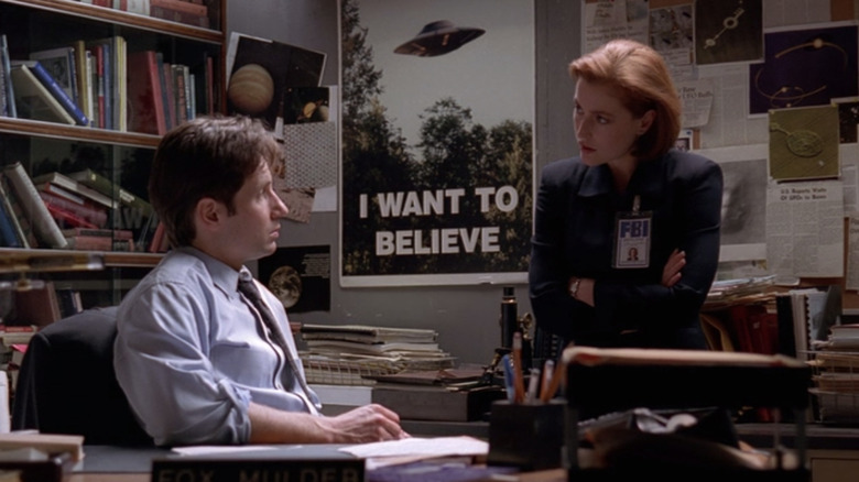 David Duchovny as Fox Mulder sitting at his desk and Gillian Anderson as Dana Sculy leaning next to an "I Want To Believe" poster in The X-Files