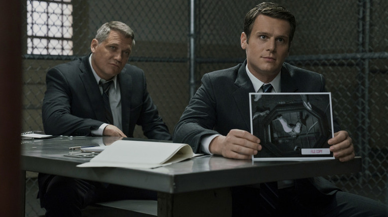 Holden Ford (Jonathan Groff) holds up a photograph with Bill Tench (Holt McCallany) seated behind him in Mindhunter