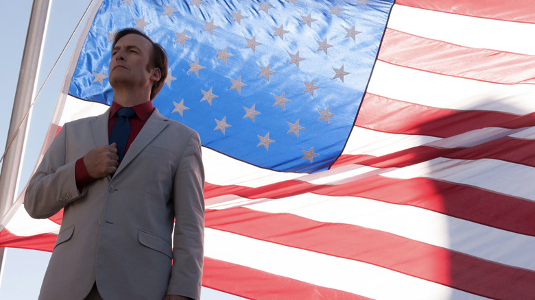 Bob Odenkirk as Jimmy McGill AKA Saul Goodman wearing a suit and standing in front of the American flag for an ad in Better Call Saul