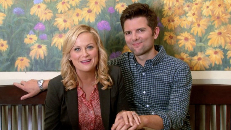 Amy Poehler as Leslie Knope and Adam Scott as Ben Wyatt sitting on the bench in front of the sunflower mural telling the camera they're engaged in Parks and Rec
