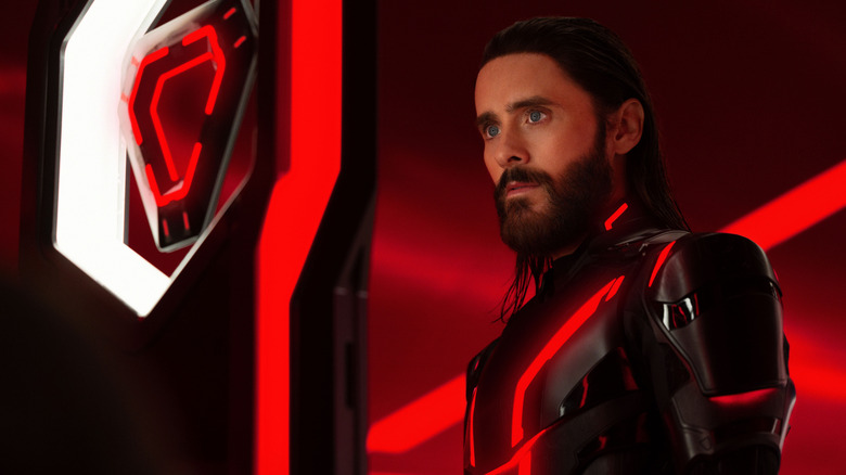 Jared Leto as Ares suited up in black armor and standing in front of a red background in Tron: Ares