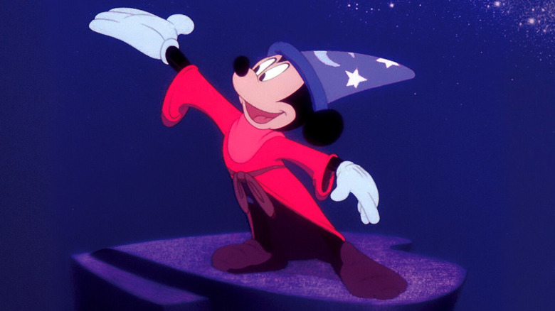 Mickey Mouse raises a hand above his head while wearing a wizard's hat in Fantasia