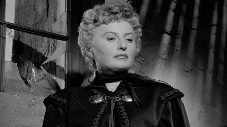 Barbara Stanwyck as Jessica Drummond looking serious in front of a broken window in Forty Guns