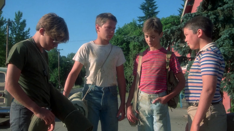 River Phoenix's Chris Chambers, Wil Wheaton's Gordie Lachance, Corey Feldman's Teddy Duchamp, and Jerry O'Connell's Vern Tessio stand on a street in a group in Stand By Me