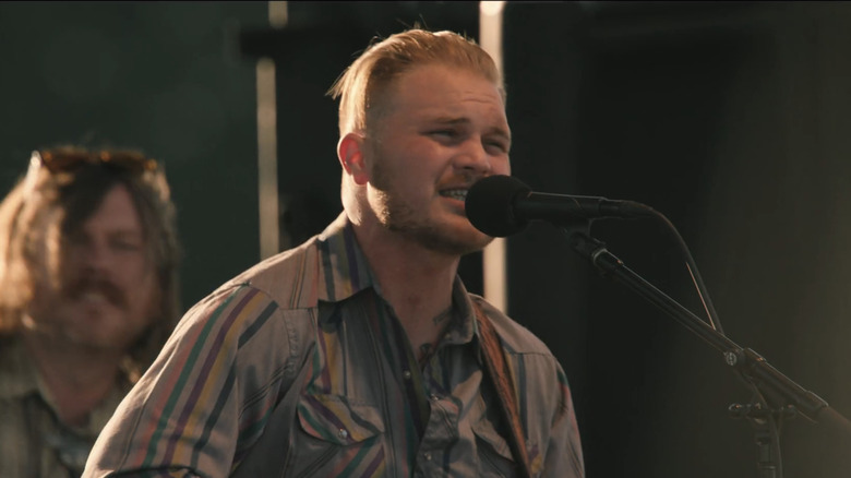 Zach Bryan sings into a microphone as he performs on-stage in Yellowstone
