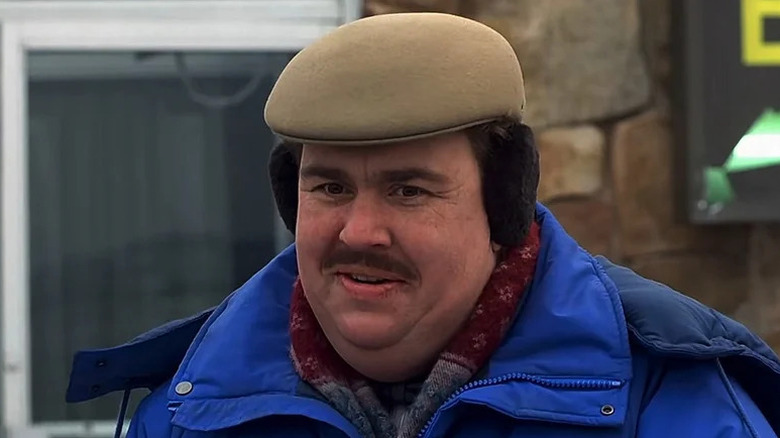 John Candy as Del, smiling in front of a hotel entrance in Planes, Trains & Automobiles