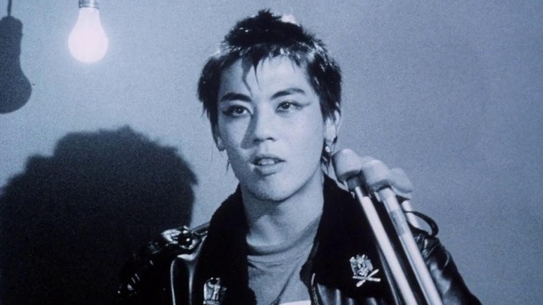 Black and white image of a young punk with crutches talking straight to camera with a lightbulb hanging creating stark shadows