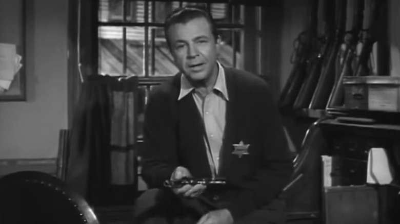 Dick Powell sits on a desk wearing a sheriff's badge in Dick Powell's Zane Grey Theatre