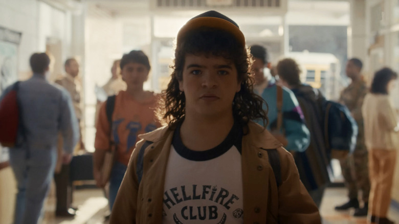 Dustin (Gaten Matarazzo) walking down a school hall in a Hellfire shirt in Stranger Things