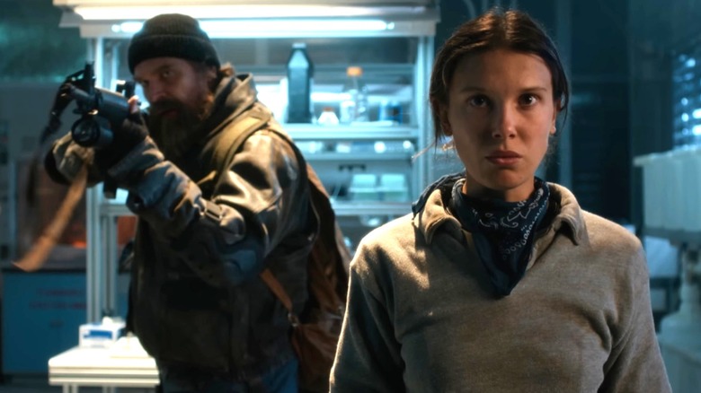Jim Hopper (David Harbour) pointing a gun and stood behind Eleven (Millie Bobby Brown) in a lab in Stranger Things