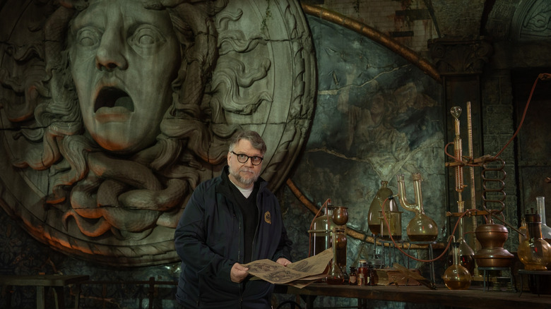 Director Guillermo del Toro standing in front of a large mural and a desk full of lab equipment in a behind-the-scenes still from the set of Frankenstein