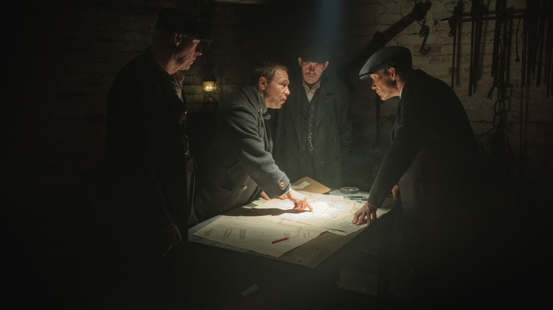Ned Dennehy as Charlie Strong, Stephen Graham as Hayden Stagg, Packy Lee as Johnny Dogs, and Cillian Murphy as Tommy Shelby standing by a table in a dark room in Peaky Blinders: The Immortal Man