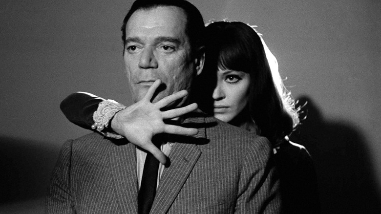 Lemmy Caution looking scared with Natacha holding a hond out from behind him in Alphaville