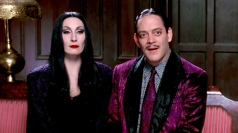 Anjelica Huston and Raul Julia sit on a couch in The Addams Family