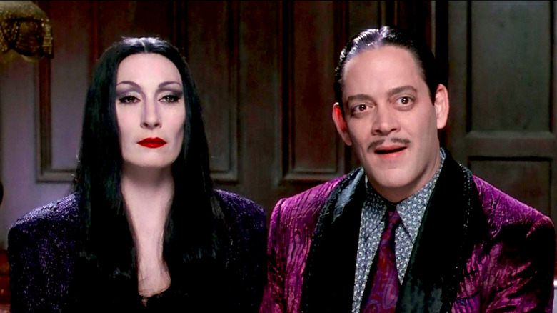 Anjelica Huston and Raul Julia sit on a couch in The Addams Family