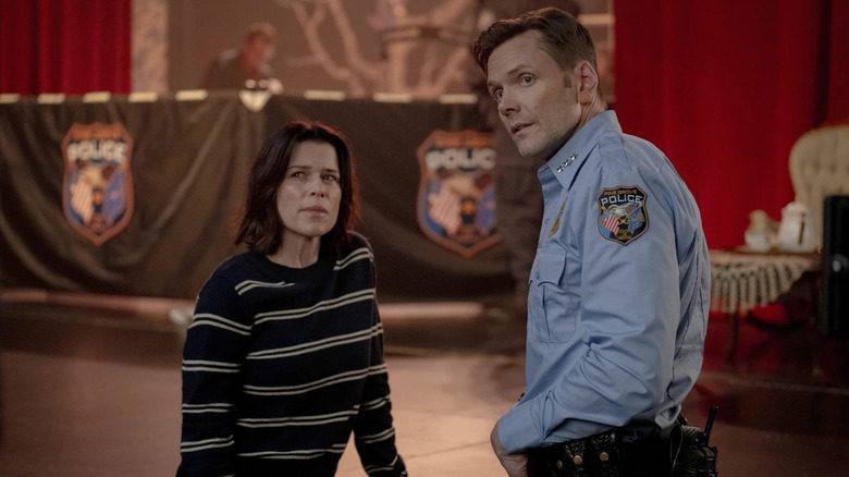 Neve Campbell as Sidney standing with Joel McHale as Mark in an auditorium in Scream 7