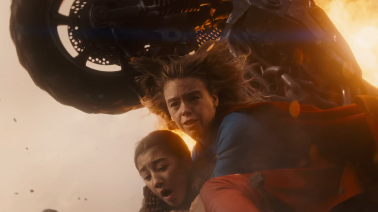 Milly Alcock as Supergirl protecting Eve Ridley as Ruthye as a vehicle flies over them in Supergirl