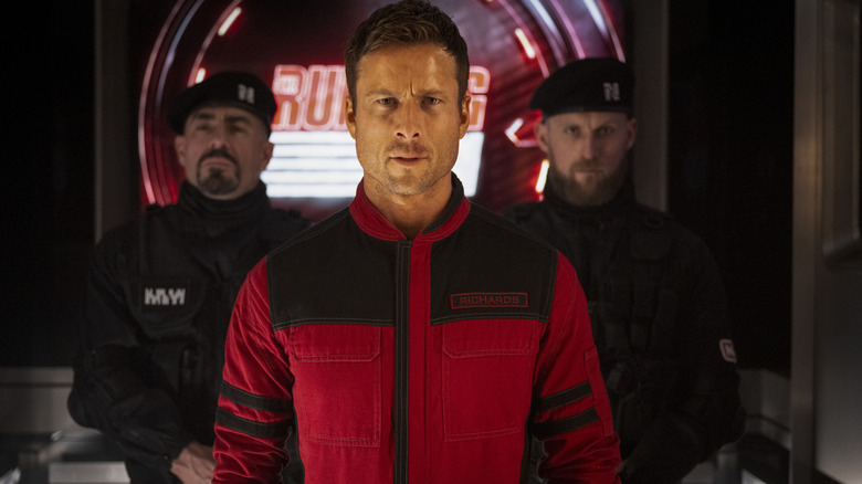 Glenn Powell as Ben Richards walking through a studio flanked by two guards in The Running Man
