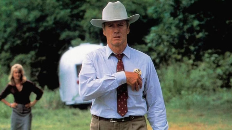 Red Garnett (Clint Eastwood) wears a serious, concerned expression as he reaches for the chest pocket beneath his police badge, while a woman stands blurred in the background with her hands on her hips in A Perfect World
