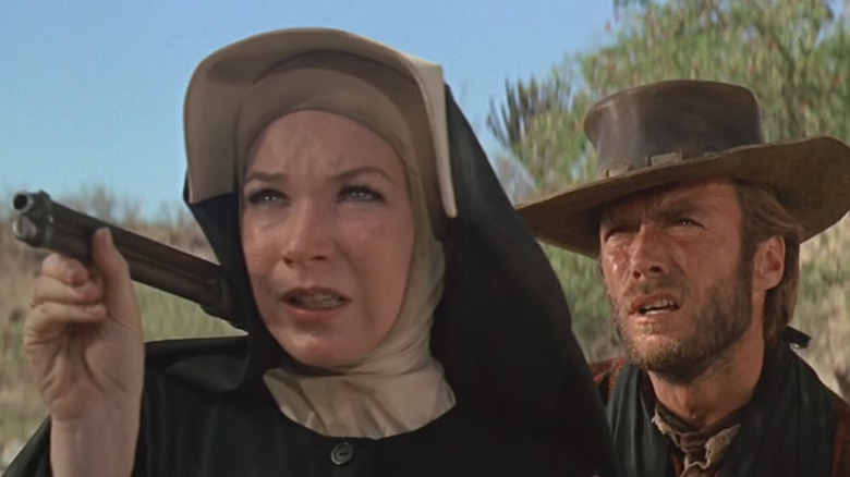 The nun Sara (Shirley MacLaine) holds a shotgun near her shoulder with a determined expression, while Hogan (Clint Eastwood), dressed in a cowboy outfit, stands behind her squinting into the distance with concern in Two Mules for Sister Sara
