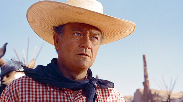 A close-up of a serious Ethan Edwards (John Wayne) wearing a large cowboy hat, red checkered shirt, and blue  neckerchief, set against the backdrop of Native American structures in The Searchers