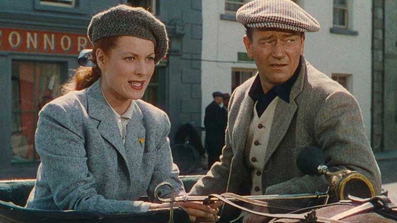 Mary Kate Danaher (Maureen O'Hara) looks on intently with her hands clasped, while Sean Thornton (John Wayne) looks off to the right side, resting his arm on the door of his 1920s-era car in The Quiet Man.