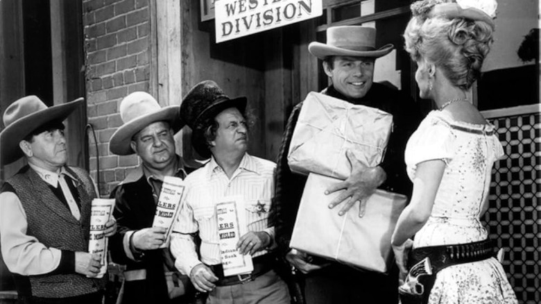 The Three Stooges in comedically large hats, standing behind Kenneth Cabot, holding two packages, as he talks to Annie Oakley. A scene from The Outlaws Is Coming.