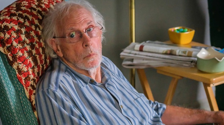 old man in striped shirt sitting on a chair with a table and newspapers next to him