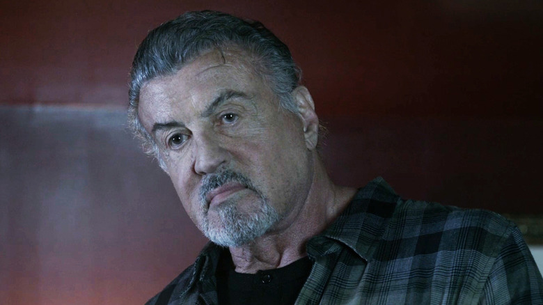 Sylvester Stallone as a Chester client stands against a red wall in AlaRom