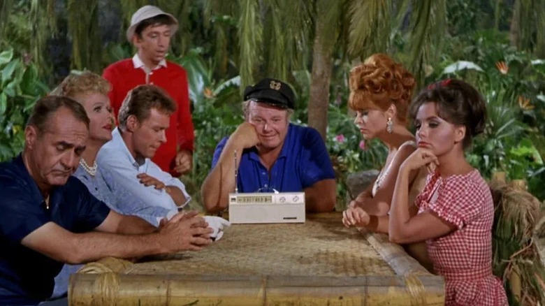 Bob Denver's Gilligan stands in the background as the rest of the Gilligan's Island cast gather around a radio in Gilligan's Island