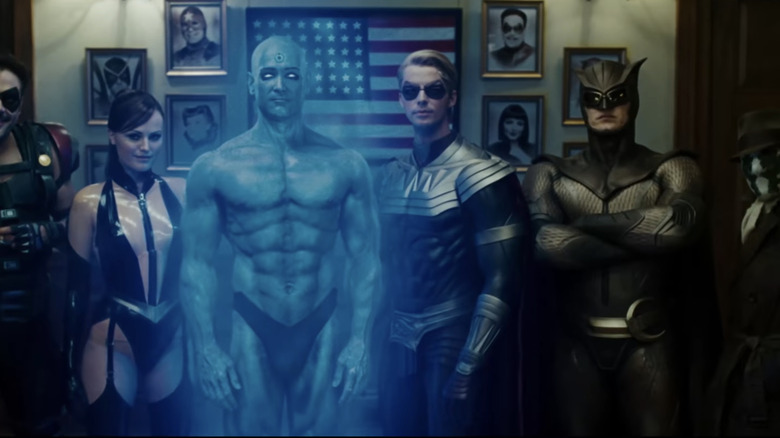 The cast of Watchmen stands in a line againts a wall with a framed US flag in Watchmen