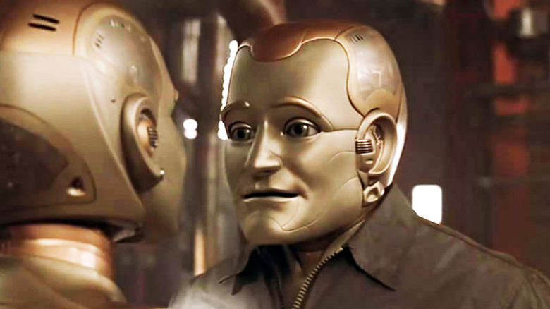 Two metallic robots face each other in a dimly lit industrial room, with a close-up of Andrew (Robin Williams) and his human-like silver and gold face with a small smile in Bicentennial Man