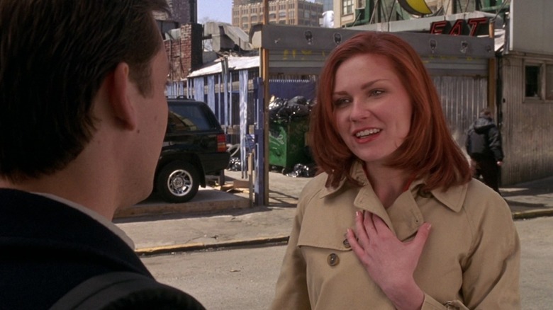 Mary Jane Watson (Kirsten Dunst) stands on a city street holding her beige trench coat closed in Spider-Man