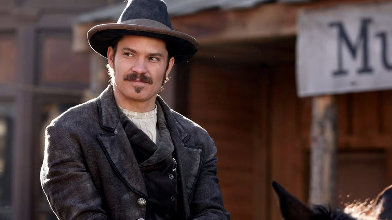 सेठ बुलॉकच्या भूमिकेत टिमोथी ऑलिफंट 1870 च्या काळातील फॅशन परिधान केलेला आणि डेडवुडमधील शहरातून घोड्यावर स्वार होत आहे
